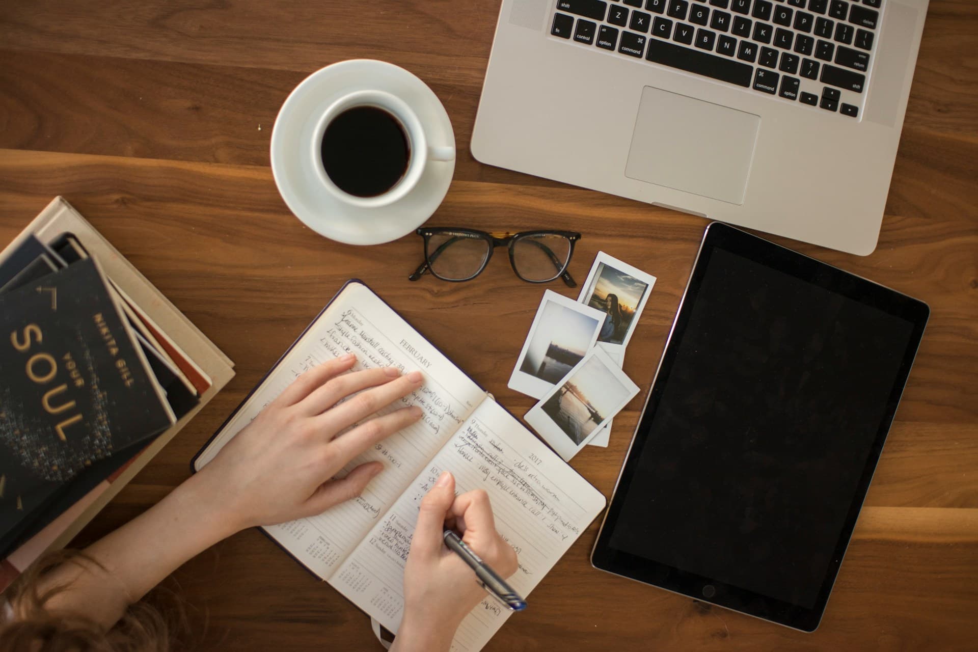 A person writes in a notebook with a laptop in front of them and a cup of coffee