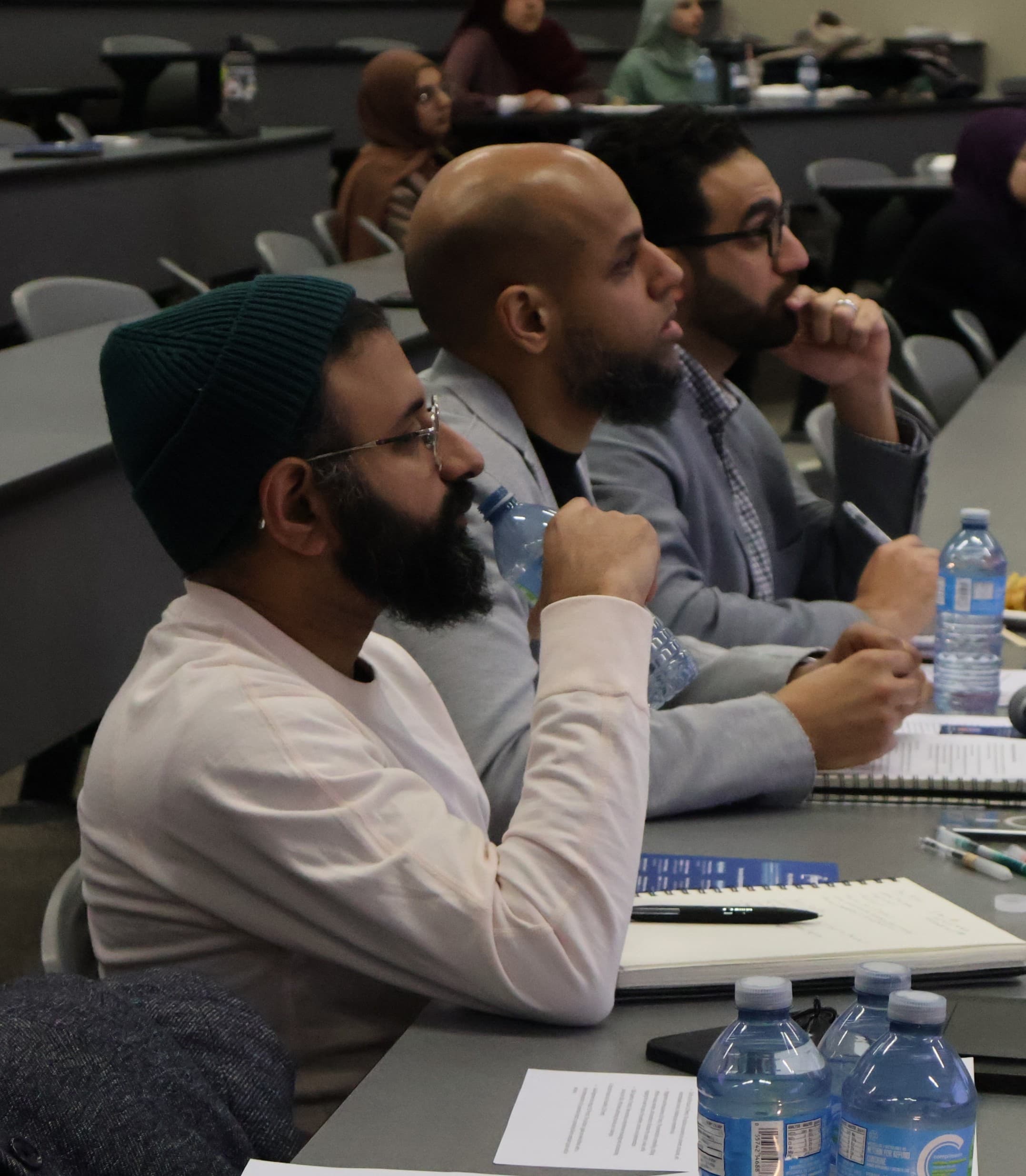 A group of men seated at a table in a lecture hall, engaged in a pitch being given by a team.