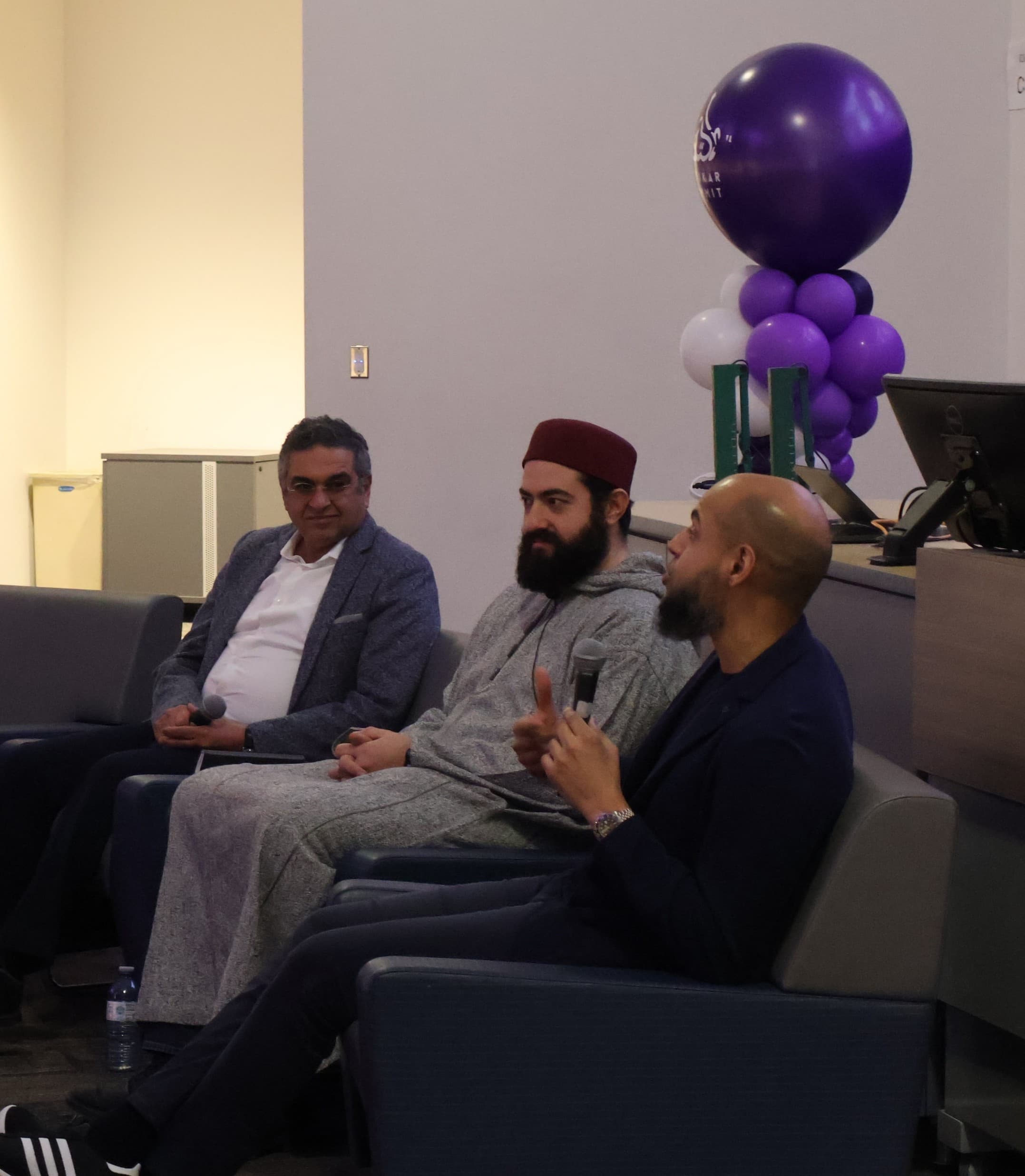Three men sitting comfortably in chairs, engaged in discussion with a larger crowd out of view.